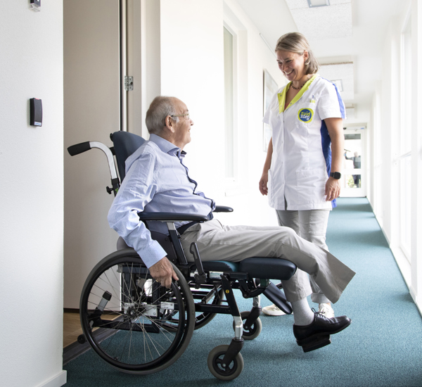 Meneer Franken in zijn rolstoel voor zijn voordeur in het appratementecomplex in gesprek met ZuidZorg verpleegkundige Renske